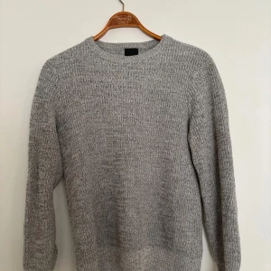Grå stickad tröja i medium - Klassisk grå stickad tröja med rund hals och långa ärmar. Tröjan har en enkel och stilren design med ribbstickade muddar vid ärmslut och nederkant. Perfekt för lager-på-lager och passar till det mesta i garderoben. Längd: 62cm, P2P: 46,5cm