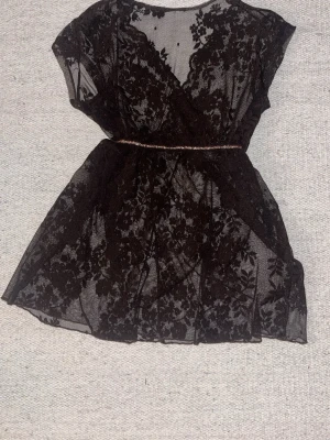 Mörkbrun spetsklänning med blommönster - Säljer en kort spetsklänning med blommigt mönster och transparent tyg. Klänningen har omlott-design och ett tunt bälte i midjan som markerar siluetten. Perfekt för dig som gillar romantiska detaljer och vill sticka ut.