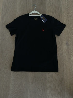 Svart t-shirt från Polo Ralph Lauren - Svart t-shirt från Polo Ralph Lauren med klassisk passform och rund hals. T-shirten har en liten röd broderad logga på bröstet och är tillverkad i mjuk bomull. Perfekt för en stilren och enkel look. Grish stockholmstil orten