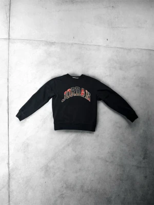 Marinblå Jordan sweatshirt med tryck - Marinblå sweatshirt från Jordan med stort broderat JORDAN-tryck i rött, vitt och svart på bröstet. Klassisk rund halsringning och ribbade muddar vid ärmslut och nederkant. Perfekt för en sportig och avslappnad stil.