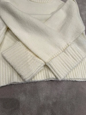 Vit stickad tröja med ribbade muddar - Klassisk vit stickad tröja med rund halsringning och breda ribbade muddar vid ärmslut och nederkant. Tröjan har en rak passform och är gjord i ett mjukt, tjockt material som känns riktigt cozy. Perfekt för kyliga dagar.