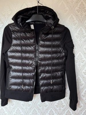 Moncler cardigan  - Snygg svart vindjacka från Moncler med quiltad framsida i glansigt material och mjuka ärmar samt rygg i bomullsmix. Loggan har gått av men GÅR ATT FIXA LÄTT. Bara att sy på! Där av de låga priset. Nypris 12,000.