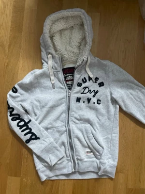 Grå Superdry hoodie med teddyfoder - Snygg ljusgrå hoodie från Superdry med dragkedja, stora fickor och broderad text på bröstet och ärmen. Huvan är fodrad med mjukt teddyfoder och har vita dragsnören. Perfekt för dig som gillar streetwear och vill ha något riktigt bekvämt.