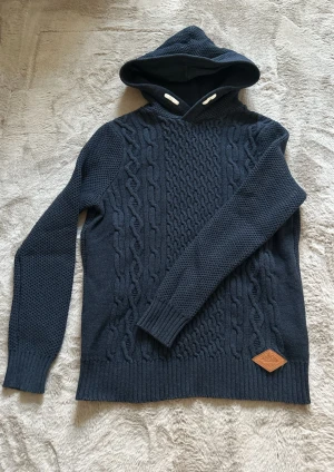 Mörkblå stickad hoodie med mönster - Säljer en snygg mörkblå stickad hoodie med kabelstickat mönster framtill och ribbade muddar. Tröjan har huva med dragsko och en liten läderpatch nertill. Perfekt för dig som gillar en avslappnad och snygg stil. Den är töjbar så den kan passa på lite större och säljer den då för att den är för liten för mig! Kom dm för mer bilder och information!