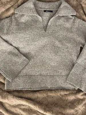 Grå stickad tröja med krage - Mysig grå stickad tröja från Gina Tricot med bred krage och v-ringad hals. Tröjan har långa ärmar och ribbade muddar vid ärmslut och nederkant. Ärmarna har dessutom ett slit längst ner. Perfekt för lager på lager och enkel att styla till olika outfits.