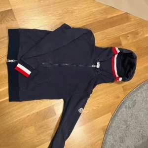 Mörkblå Moncler hoodie med dragkedja - Säljer en original Moncler hoodie i marinblå färg, storlek S. Klassisk zip-up modell med huva och dragkedja framtill. Stilrena röd-vita detaljer på huvan och ärmslut som ger ett sportigt och exklusivt intryck. Hoodien är i bra skick men har ett litet hål, enkelt att laga eller sy igen och som inte påverkar användningen nämnvärt. I övrigt inga större fläckar eller skador. Bekväm och varm, perfekt till vardags eller streetwear. 📏 Storlek: S 🎨 Färg: Marinblå 👕 📦 Skickas snabbt