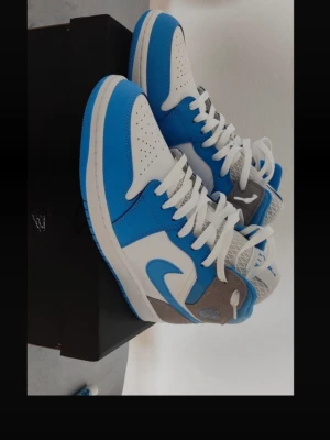 Nike Air Jordan 1 Mid blå/vit/grå - ‼️Priset är flexibelt‼️                                    Snygga Air Jordan 1 Mid sneakers från Nike i blått, vitt och grått. Skorna har klassisk hög siluett, vita snören och ikoniska swoosh-loggan på sidan. Ovandelen är i läder och textil med perforerad tå för extra ventilation. Perfekta för dig som gillar streetwear.