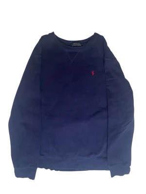 Mörkblå sweatshirt från Polo Ralph Lauren - Mörkblå sweatshirt från Polo Ralph Lauren med klassisk rund halsringning och röd broderad logga på bröstet. Tröjan har långa ärmar och ribbade muddar vid ärmslut och nederkant. Perfekt för en avslappnad och stilren look.Obs de finns en liten skada på ena armen som visas på bilden