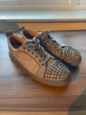 Louboutin grå - Säljer ett par gråa sneakers från Christian Louboutin i mocka med nitar och med den klassiska röda sulan. St.44 men tycker personligen att de är lite mindre i storleken.  Kommer med vad som ses på den sista bilden, bland annat kartong, dustbag m.m. Osäker på äktheten så kontakta för mer bilder om så önskas. Kan även tänka mig att droppa priset om det går smidigt😊