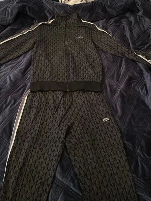 Lacoste tracuit storlek S - Bilderna visar ett svart mönstrat träningsset från Lacoste med dragkedja och vita detaljer, syns på bilderna.