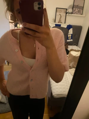 Ljusrosa stickad kofta med knappar - Säljer en ljusrosa stickad kofta med korta ärmar och v-ringad hals. Koftan har ribbad struktur och knäpps med flera vita knappar framtill. Perfekt för dig som gillar mjuka och mysiga plagg med lite söt vibe.