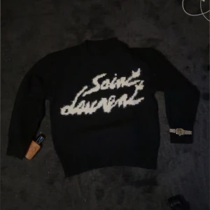 Svart stickad Saint Laurent tröja - Fick denna nu i julklapp men tyvärr så passar den inte riktigt…Svart stickad tröja från Saint Laurent med vit, fluffig logotyp framtill. Rund hals och långa ärmar. Tröjan har en loose passform och är perfekt för dig som vill ha en statement piece med lyxig vibe. Materialet är mjukt och skönt mot huden.