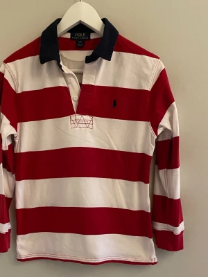 Randig långärmad t-shirt Polo Ralph Lauren - Långärmad piké i röd och vita ränder. Den har en mörkblå krage och är i storlek 10-12 år. Säljer då den inte passar längre. Hör av er vi funderingar