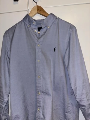 Ljusblå skjorta från Ralph Lauren - Snygg ljusblå skjorta från Ralph Lauren med klassisk button-down krage och broderad logga på bröstet. Skjortan har långa ärmar, normal passform och är tillverkad i mjuk bomull. Perfekt för dig som gillar stilrena och tidlösa plagg. 