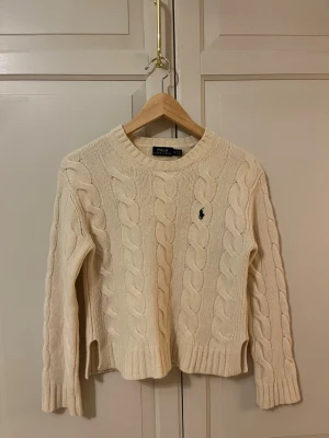 Beige kabelstickad tröja Polo Ralph Lauren - köpt i stockholm, fått som present men passar tyvärr inte, jätte fint skick, cremefärgad med mörkblått märke, slits på sidorna, vid frågor eller önskemål av bilder är det bara att skriva, skickar inom en dag! 💕. Tröjan har rund hals, ribbade muddar och ett broderat mörkblått Polo-logo på bröstet. Den är långärmad och har en klassisk, lite tightare passform med slitsar i sidorna.