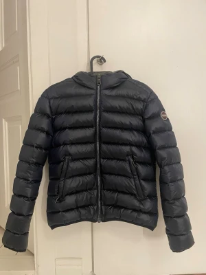 Svart pufferjacka från Colmar - Säljer en snygg mörkblå pufferjacka från Colmar med huva och två fickor med dragkedja. Jackan har ett stilrent, quiltat mönster och logotyp på ärmen. Perfekt för kalla dagar och har en snygg, modern look som funkar till streetwear. Jackan är som ny och är bara använd några gånger.💕Hör av er om ni undrar något eller om ni vill ha fler bilder Jackan är i storlek XS/S.