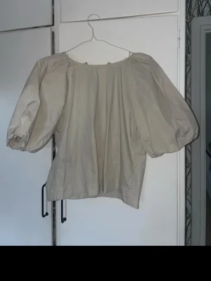 Beige blus med puffärmar från H&M - Säljer en beige blus från H&M i storlek M. Blusen har bred halsringning och stora puffärmar som ger en trendig siluett. Tillverkad i ett mjukt bomullsmaterial och har en loose passform. Perfekt att styla med jeans eller kjol för en cool look.