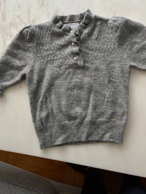 Grå glittrig stickad tröja Holly & Whyte - Snygg grå stickad tröja från Holly & Whyte med glittriga inslag och pärlknappar framtill. Tröjan har korta ärmar, ribbad kant och ett diskret mönster upptill. Perfekt för dig som gillar detaljer och vill ha något extra i garderoben.