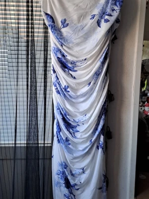 Vit långklänning med blå blommor - Säljer en vit långklänning med tunna axelband och blått blommigt mönster. Klänningen har draperad design längs hela längden som ger en snygg och unik look. Materialet känns mjukt och lätt, perfekt för varmare dagar.