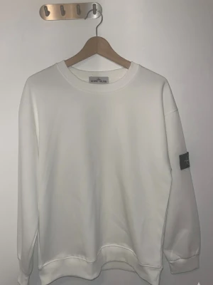 Vit sweatshirt från Stone Island - Vit sweatshirt från Stone Island med klassisk rund hals och ribbade muddar. Tröjan har lång ärm och den ikoniska Stone Island-loggan på vänster ärm. Materialet är mjuk bomull som känns skön mot huden. Perfekt för dig som gillar stilrena och enkla plagg. Har aldrig använts då den är stör för mig. Den har legat i garderoben i flera månader och jag vill bli av med den