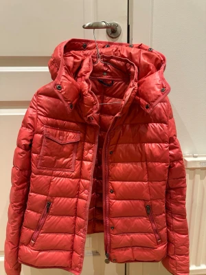 Röd pufferjacka från Moncler - Säljer en korall röd pufferjacka från Moncler med huva och snygga quiltade paneler. Jackan har dragkedja framtill, två fickor med dragkedja och en bröstficka med knapp. Moncler-logga på ärmen och coolt serietryck på insidan. Perfekt för kalla dagar!