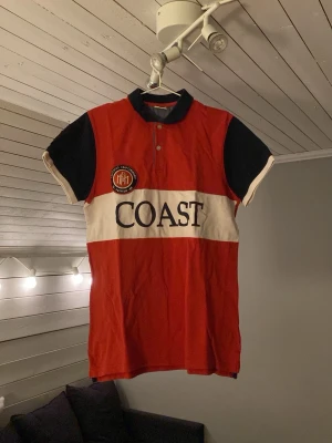 Röd och svart Coast t-shirt med tryck - Säljer en snygg t-shirt från Coast med röd kropp, svarta ärmar och krage samt vit panel med stort tryck. T-shirten har korta ärmar, knappslå och ett broderat märke på bröstet. Perfekt för dig som gillar sportig och stilren design.