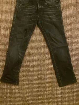 Replay Anbass svarta jeans, stl 31 - Svarta Replay Anbass jeans med snygga slitningar och klassisk femficksdesign. Jeansen har en smal passform och är tillverkade i stretchigt denim för extra komfort. Märkespatch bak i midjan och coola detaljer på bakfickorna. Perfekt för dig som gillar streetstyle.