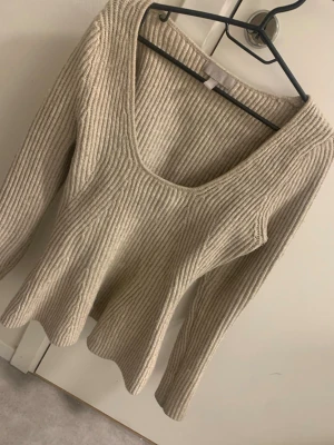 Beige ribbad stickad tröja med v-ringning - Säljer en beige stickad tröja med ribbad struktur och snygg v-ringad hals. Tröjan har lång ärm och sitter figurnära upptill med en lätt utsvängd nederdel. Perfekt för dig som gillar enkel och stilren design med lite extra form.