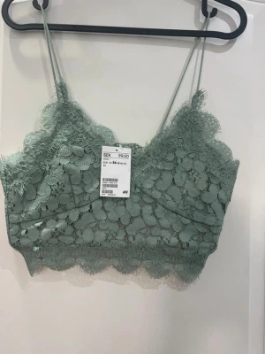 Grön spetsig linnetopp från H&M - Säljer en ljusgrön linnetopp från H&M med tunna axelband och vacker spets över hela toppen. Toppen har blommigt spetsmönster, croppad passform och en diskret dragkedja i sidan för enkel påklädning. 
