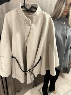Beige cape-kappa med bälte - Snygg beige cape-kappa från M Collection med hög krage och dekorativ silverknapp vid halsen. Kappan har ett svart bälte i midjan som ger en cool touch och ett flowy fall. Perfekt för dig som vill ha en stilren och trendig look.