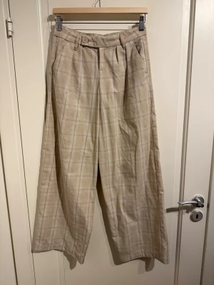 Rutiga vida kostymbyxor  - Beiga byxor med låg midja. Materialet är bomull.