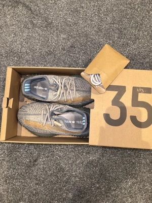 Adidas Yeezy Boost 350 V2 grå/blå - Säljer ett par Adidas Yeezy Boost 350 V2 sneakers i grått och blått med beige detaljer. Skorna har stickad ovandel, snörning och en platt sula med tydlig struktur. Ikonisk Yeezy-design med mönstrad mesh och Boost-dämpning för extra komfort. Skorna kommer med ett extra par helt oanvända skosnören och låda. Skorna går även att tvätta i tvättmaskinen. Glömm inte att det är ORGINAL ( äkta )