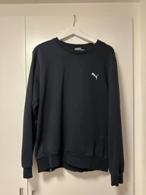 Svart sweatshirt från Puma, storlek M - Svart sweatshirt från Puma i storlek M. Tröjan har rund hals, ribbade muddar och ett litet Puma-logo broderat på bröstet. Tröjan är i fint skick men färgen är lite uttvättad. 