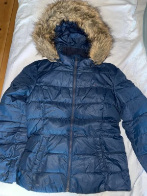 Mörkblå pufferjacka från Tommy Hilfiger - Säljer en mörkblå pufferjacka från Tommy Hilfiger med huva och fejkpäls. Jackan har dragkedja framtill och två fickor på sidorna. Perfekt för kalla dagar med sin fluffiga look och klassiska design. Liten logga på bröstet. Storlek 164