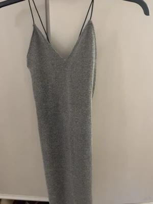 Glittrig silver klänning XXS - Säljer en snygg långklänning i glittrigt silvermaterial med smala svarta axelband och v-ringad hals. Klänningen är ärmlös och har en stilren, figurnära passform som ger en cool och trendig look. Perfekt för dig som gillar att sticka ut med lite extra glitter.