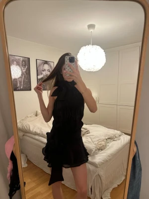 Svart glittrig klänning med rosetter - Supersnygg svart kort klänning från Gina Tricot med stor rosett vid halsen och midjan. Rosetterna går att justera. Klänningen har volangdetaljer nertill och är ärmlös med en elegant, glansig finish. Perfekt för fester eller middagar🤩🤩