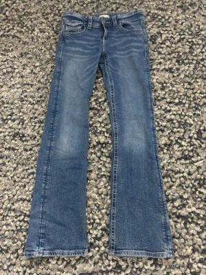 Blå bootcut jeans från Gina Tricot - Snygga blå jeans från Gina Tricot i storlek 158. Modellen är bootcut med klassisk femficksdesign och coola detaljer med paljetter och broderi på bakfickorna. Jeansen har normal passform och är tillverkade i jeansmaterial med dragkedja och knapp framtill.