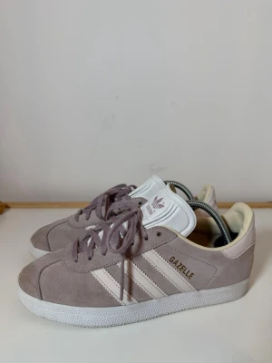 Adidas Gazelle sneakers i ljuslila mocka - Säljer ett par Adidas Gazelle sneakers i ljuslila mocka med vita detaljer och klassiska tre ränder på sidan. Skorna har snörning, vit platt sula och rund tå. Adidas-logga i lila på hälen och tungan. Perfekt för dig som gillar retrostil och snygga färgkombinationer.
