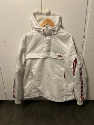 Alpha Industries Jacka - Snygg vit vindjacka från Alpha Industries i storlek XL. Jackan har huva, röda detaljer och dragkedjor. I gott skick!