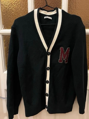 Svart cardigan  - Svart stickad cardigan från Zara i storlek M. På bröstet sitter ett stort vinrött M med grå kant. Perfekt för dig som gillar college-vibe och vill ha något snyggt och bekvämt.