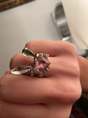 Söt kaninring med rosa hjärta - Unik ring i silverfärgad metall med kaninöron och ett stort rosa hjärtformat glas i mitten. Ringen har små klara stenar runt hjärtat och en lekfull design som verkligen sticker ut. Perfekt accessoar för dig som gillar gulliga detaljer och statement-smycken.