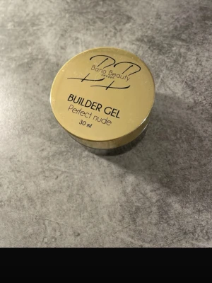 Bano Beauty Builder Gel Perfect Nude - Builder Gel Perfect Nude från Bano Beauty i en rund burk på 30 ml. Gelen har en mjuk nude-rosa färg och används för att bygga och förstärka naglar. Förpackningen är guldig med svart text och har en praktisk skruvlock. Perfekt för dig som gillar att fixa naglarna hemma.