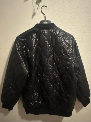 Svart quiltad bomberjacka från Yut - Quiltad svart bomberjacka från Yut med glansig yta och dragkedja framtill. Jackan har ribbade muddar vid ärmslut och nederkant, samt ett snyggt mönster i quiltad stil. Perfekt för dig som gillar streetwear och vill ha en jacka med cool vibe.