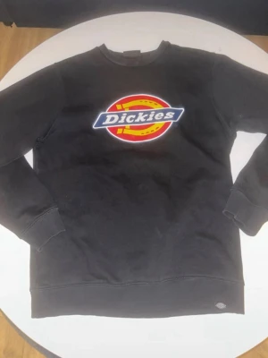 Svart sweatshirt från Dickies, S - Svart sweatshirt från Dickies i storlek small med stort färgglatt logotryck på bröstet. Tröjan har rund hals, långa ärmar och ribbade muddar vid ärmslut och nederkant. Mjuk insida i bomullsmix som gör den skön att ha på sig.