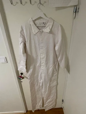 Vit byxdress med krage, storlek S - Säljer en vit byxdress i storlek S med klassisk krage och lång ärm. Byxdressen har raka ben och en slits framtill, samt fickor på sidorna. Materialet känns som bomull och plagget har en stilren, minimalistisk look.