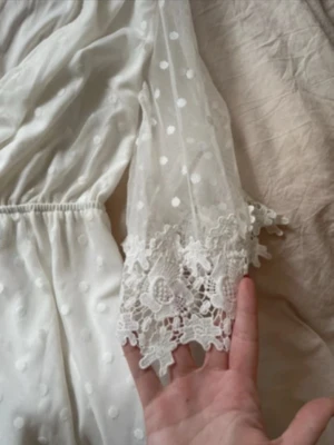 Vit prickig byxdress från Parisian - Säljer en vit byxdress från Parisian med prickigt mönster och snygga spetsdetaljer vid ärmslut och benslut. Modellen har omlottliv, transparenta ärmar och knyt i midjan. Perfekt för dig som gillar romantisk och feminin stil.