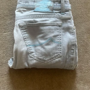 Ljusgrå jeansbyxor från Jacob Cohen - Snygga ljusgrå jeansbyxor från Jacob Cohen med en klassisk femficksdesign. Byxorna har en slim fit passform. Ostryckna på bilderna men kommer såklart stryka dem innan köp. Det är det påväg till ett hål på bild 3 annars så är skicket bra. Storlek är 34/34 och sitter true to size