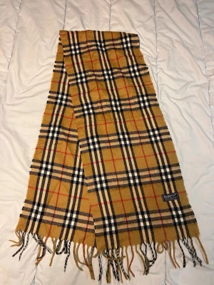 Rutig Burberrys halsduk i ull - Säljer en klassisk rutig halsduk från Burberrys i 100% lammull. Färgerna är beige, svart, vit och röd med typiskt rutmönster och fransar i ändarna. Märkt med Burberrys-tag. Perfekt accessoar för att lyfta din outfit. N14