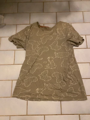 Beige t-shirt med kartmönster - Säljer en beige t-shirt med coolt kartmönster över hela plagget. T-shirten har rund hals och korta ärmar, perfekt för dig som gillar unika prints. Materialet känns mjukt och skönt, troligtvis bomull. Passar dig som vill sticka ut med en annorlunda look.