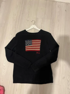 Marinblå stickad tröja från Ralph Lauren - Marinblå stickad tröja från Ralph Lauren med amerikansk flagga i rött, vitt och blått på bröstet. RL-broderi under flaggan. Rund hals och långa ärmar med ribbade muddar och nederkant. Perfekt för dig som gillar klassisk och cool stil.